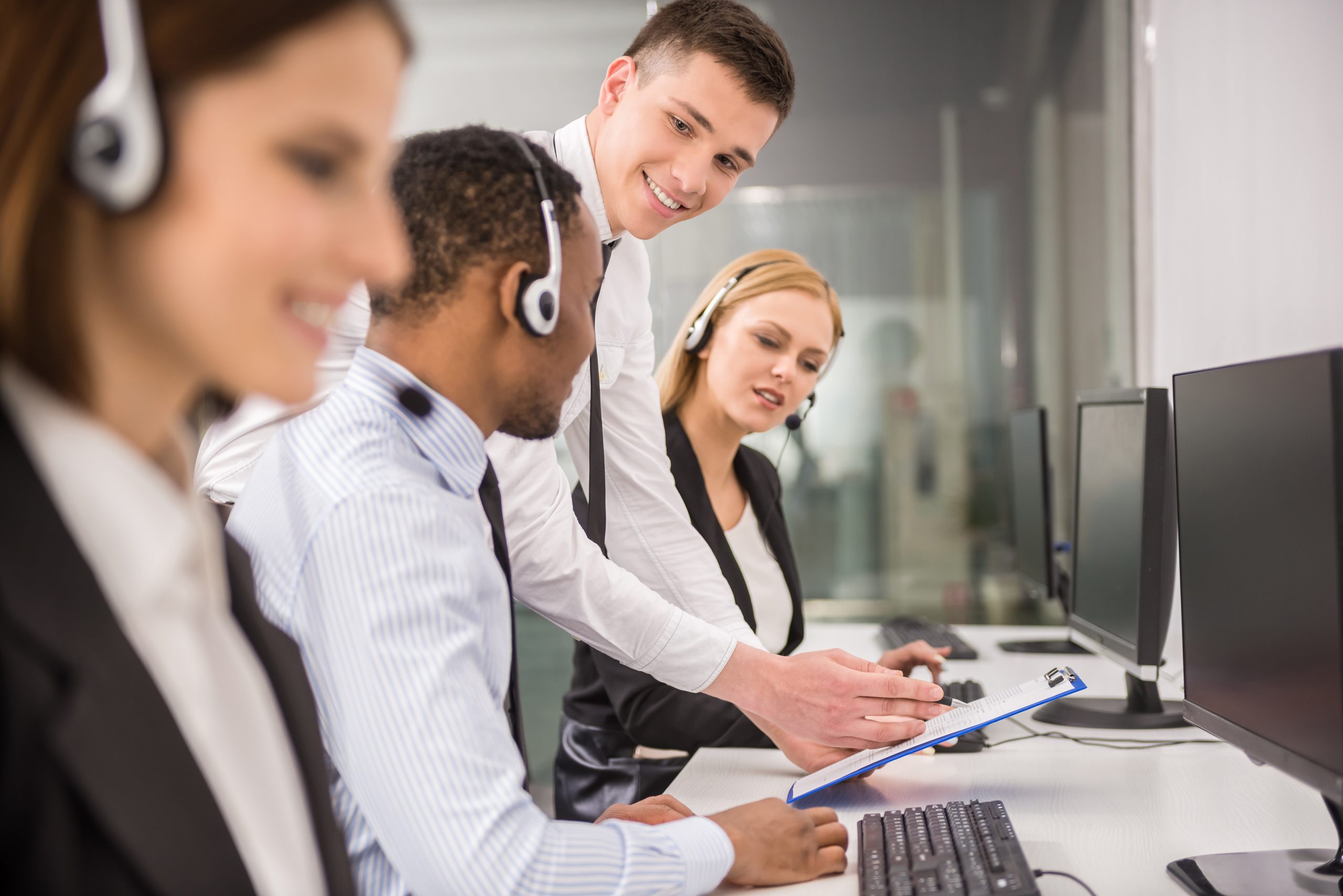 Young white male manager with clip board in hand, training in other staff member in front of computers